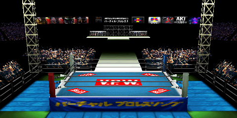 Screenshot of Dome Road, focused on the
ring area. A few items in the far distance: box arts of four of AKI's US-released
N64 wrestling games on the left side; a display in the center featuring a clock
and some text, as well as the AKI Corporation logo; some further logos on the right
hand side. A stage can be seen in the upper center. This leads to the ring area,
though an unlit area separates the two. Some fans are ringside, with light trusses
near the corners. The ring mat design has five 'V.P.W.' logos, each consisting of
a red rectangle and white text. Most of the non-logo portion is teal, but there is
a white border section at the edges. The ring skirt is a darker blue, with yellow
Japanese text. Like Budokan, the outside corner areas without mats are green.
The outside mats are light blue, similar to the ones in AKI Hall.