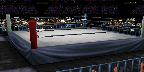 Screenshot of 'USA Dome', focused on
the ring area. Many fans appear in the distance, representing the far seats.
There is also a contingent of fans closer to the ring. Blue guardrails separate
the ringside fans from the action. The ring itself appears on an angle. The primary
mat design is a gray square, bordered by a darker gray edge. This dark gray continues
to the ring skirt design. Outside the ring are four sets of black mats, with the
corners being a lighter blue-ish concrete.